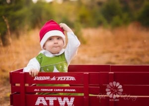 A professional image of a toddler in a radio flyer with a Santa hat on.
