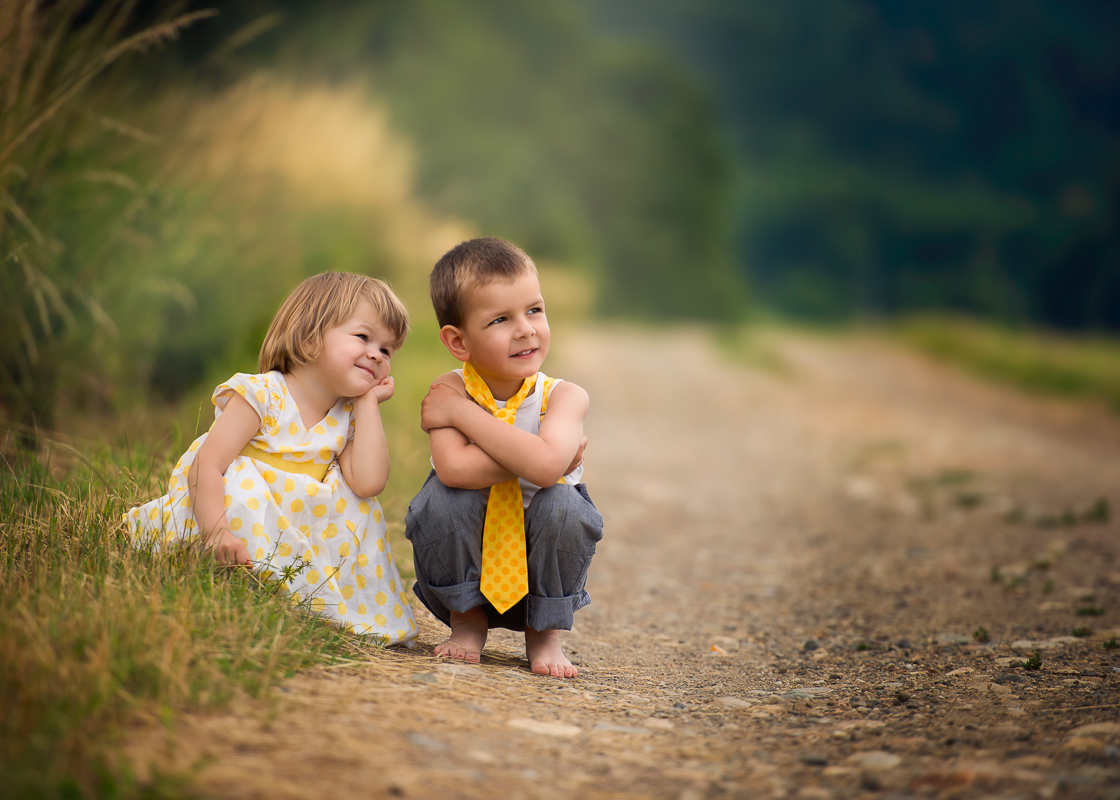Two adorable children (boy and girl) crouching down in a middle of a dirt road.