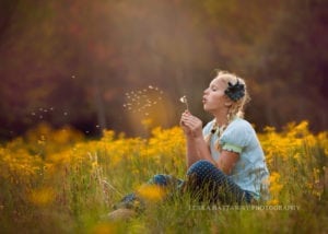 A colorful image of a girl sitting among many flowers and blowing dandelion.