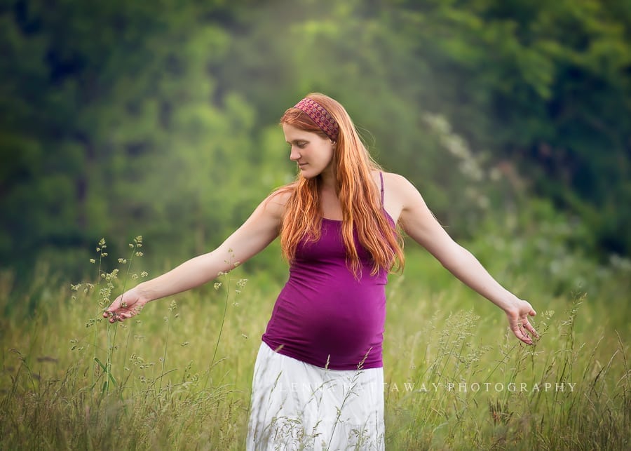 An image of pregnant woman spreading her arms touching grass around her. The mom has long red hair.