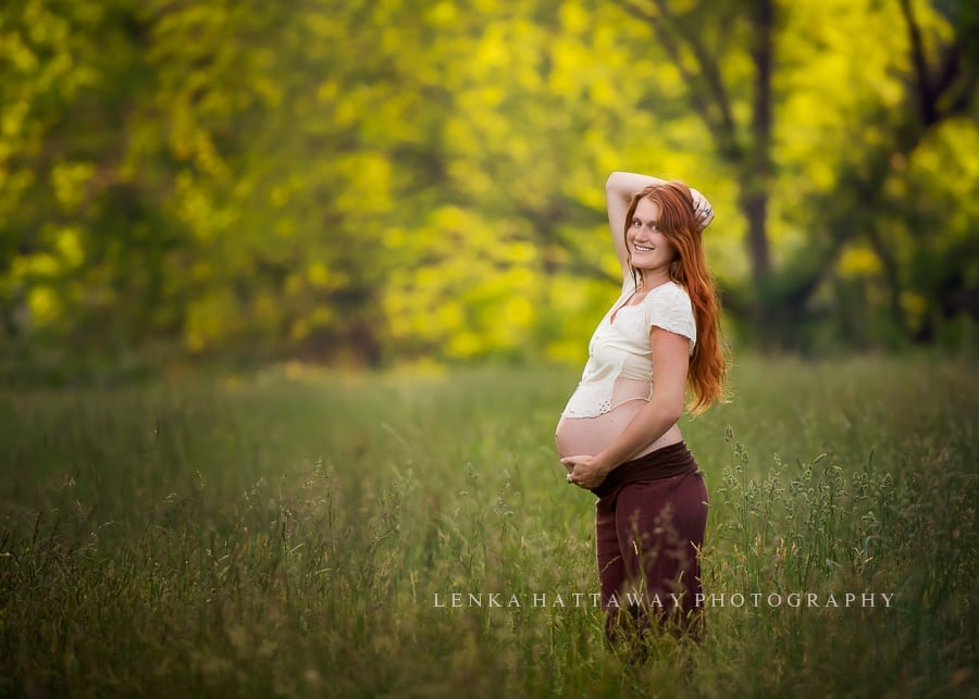 A pregnant mom with long red hair standing in grass with one arm up over her head.