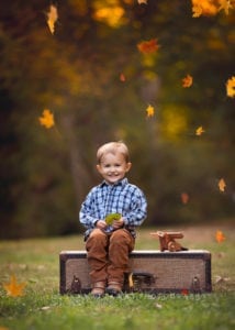 A photo of a cute little boy sitting on a trunk with leaves falling around him. Photo taken at the botanical Gardens of Asheville.