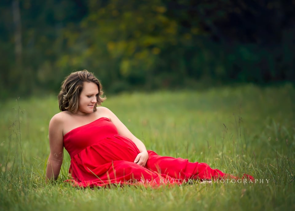 A maternity shot of a mama in a red gown sitting down and looking at her belly.