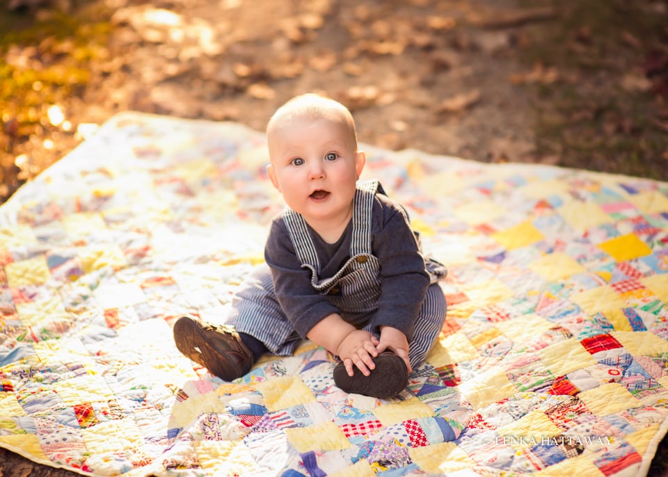 A cute baby sitting on a blanket and looking up into the camera.