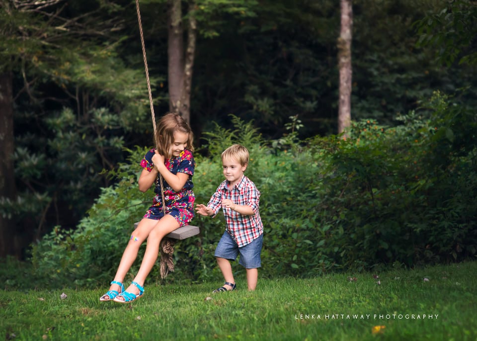 A photo of two children. One child is swinging, teh other is pushing teh swing.