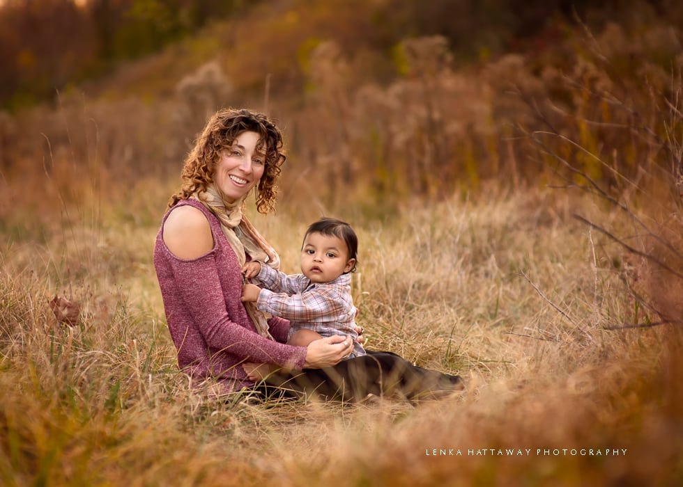 Mom holding a baby boy on her lap, both are sitting down in grass.