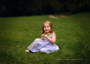 A precious little girl sitting on grass and looking into the camera.