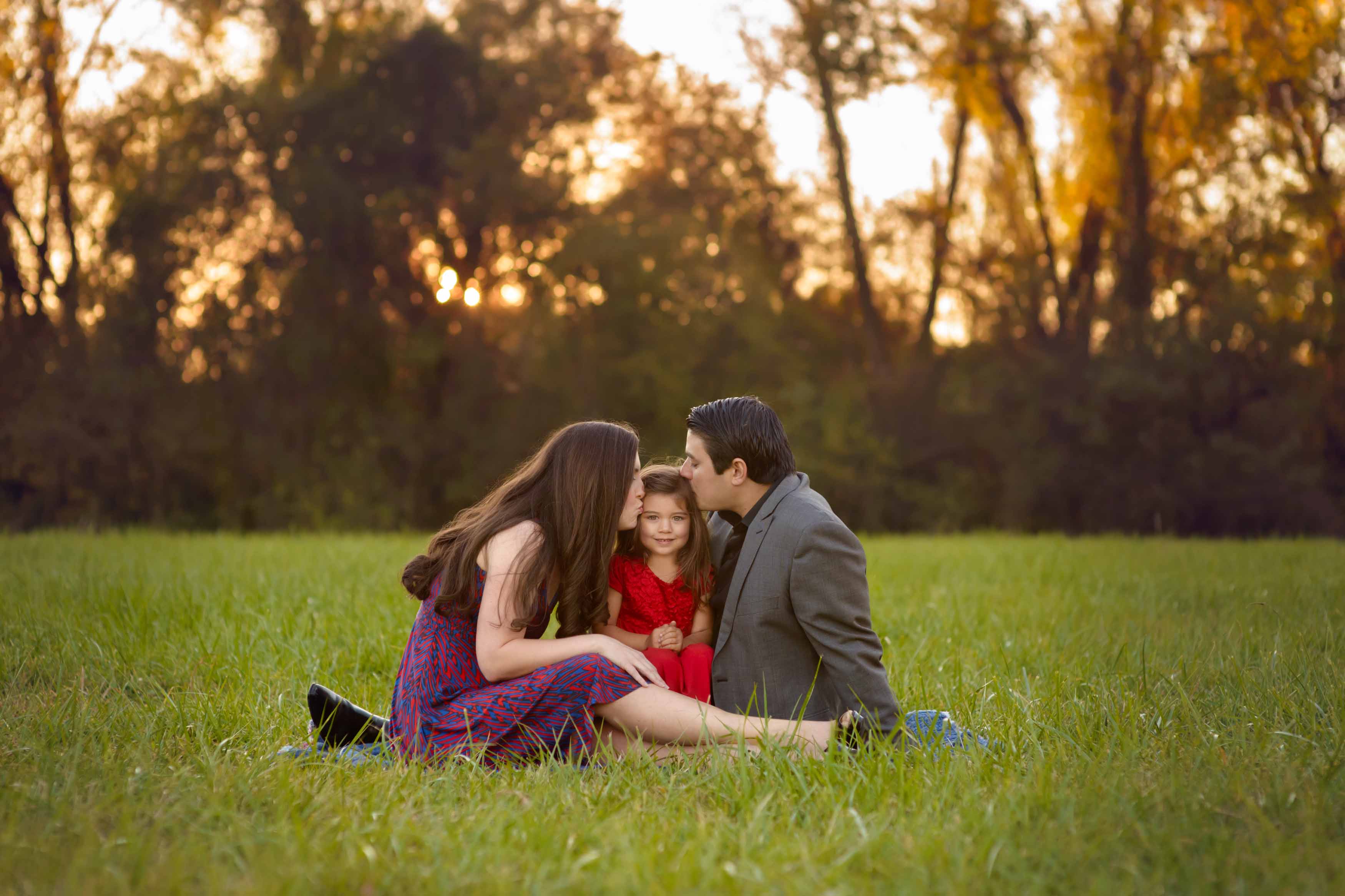 Parents kissing their child while sitting in the grass.