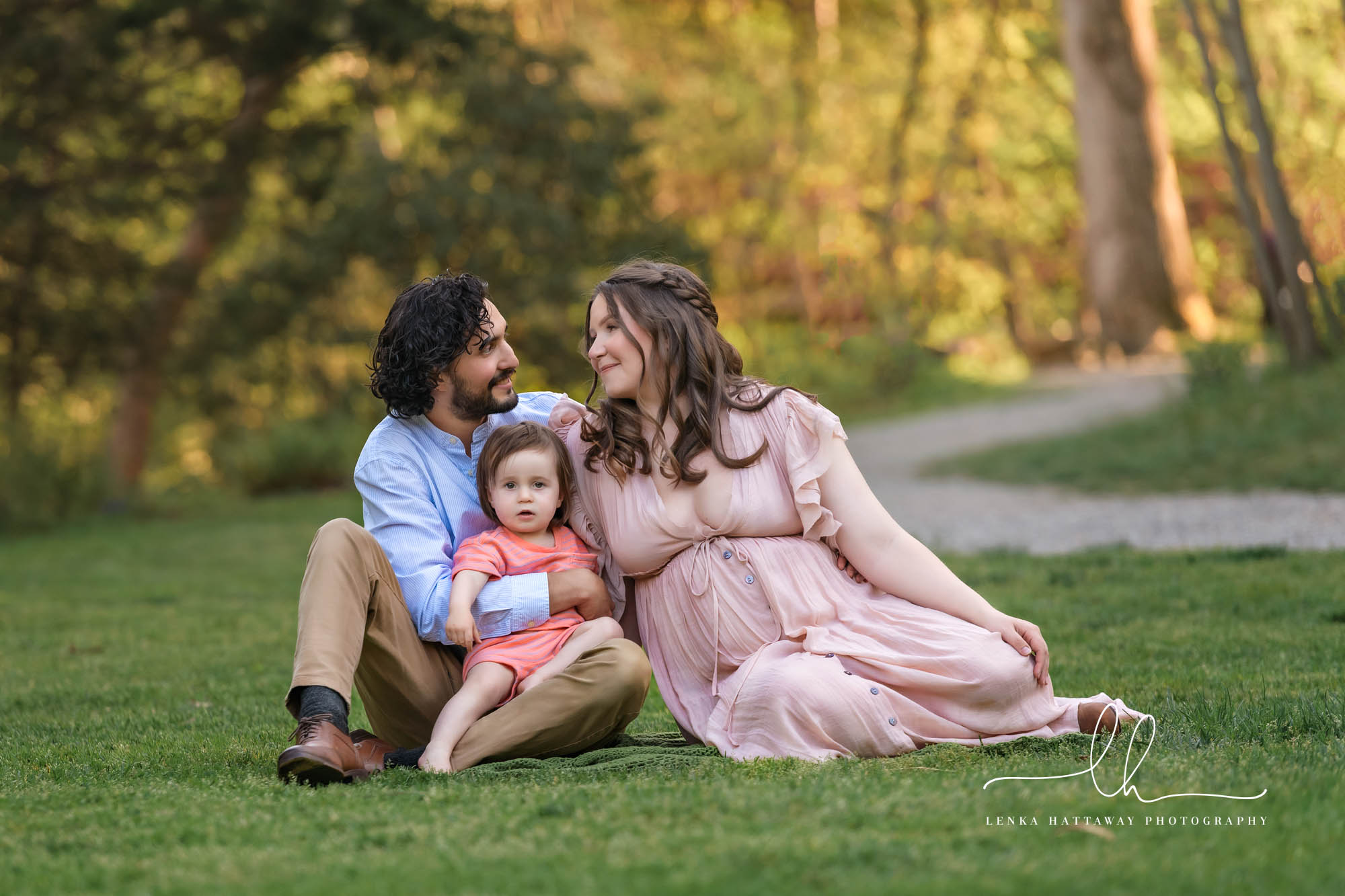 Family of three expecting another baby sitting on grass during their maternity photo session.