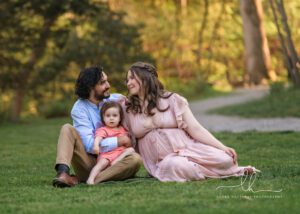 Family of three expecting another baby sitting on grass during their maternity photo session.